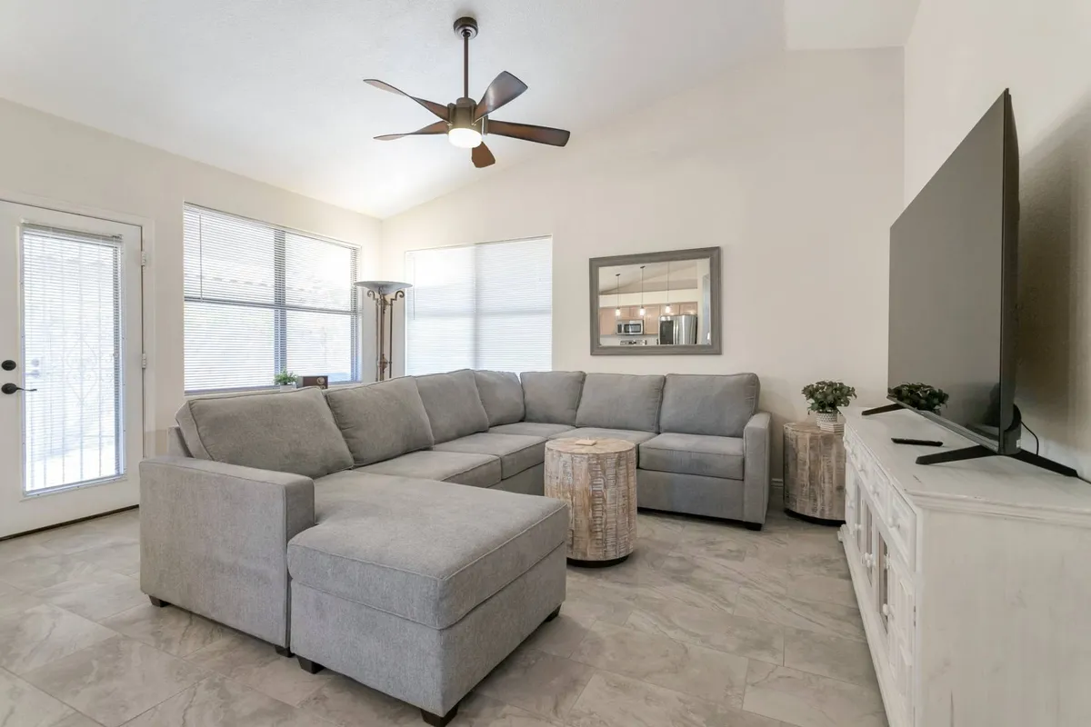 A welcoming interior living space featuring a large sectional sofa facing a television, surrounded by wooden side tables and windows that let in natural light. The room is designed for comfort and group entertainment, ideal for catching game highlights or enjoying downtime.