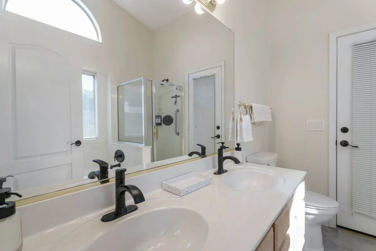 A modern bathroom with double sinks, a large mirror, a glass-enclosed shower, and contemporary light fixtures. The space is bright, fresh, and well-equipped for morning or evening routines.