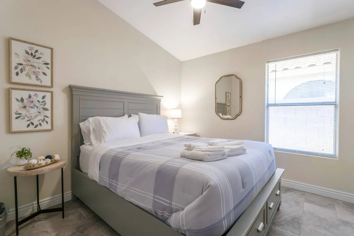 A tranquil bedroom with a neatly made bed, minimalist decorative art, and a mirror, bathed in gentle natural light. The serene atmosphere offers a sense of calm, privacy, and restful escape.
