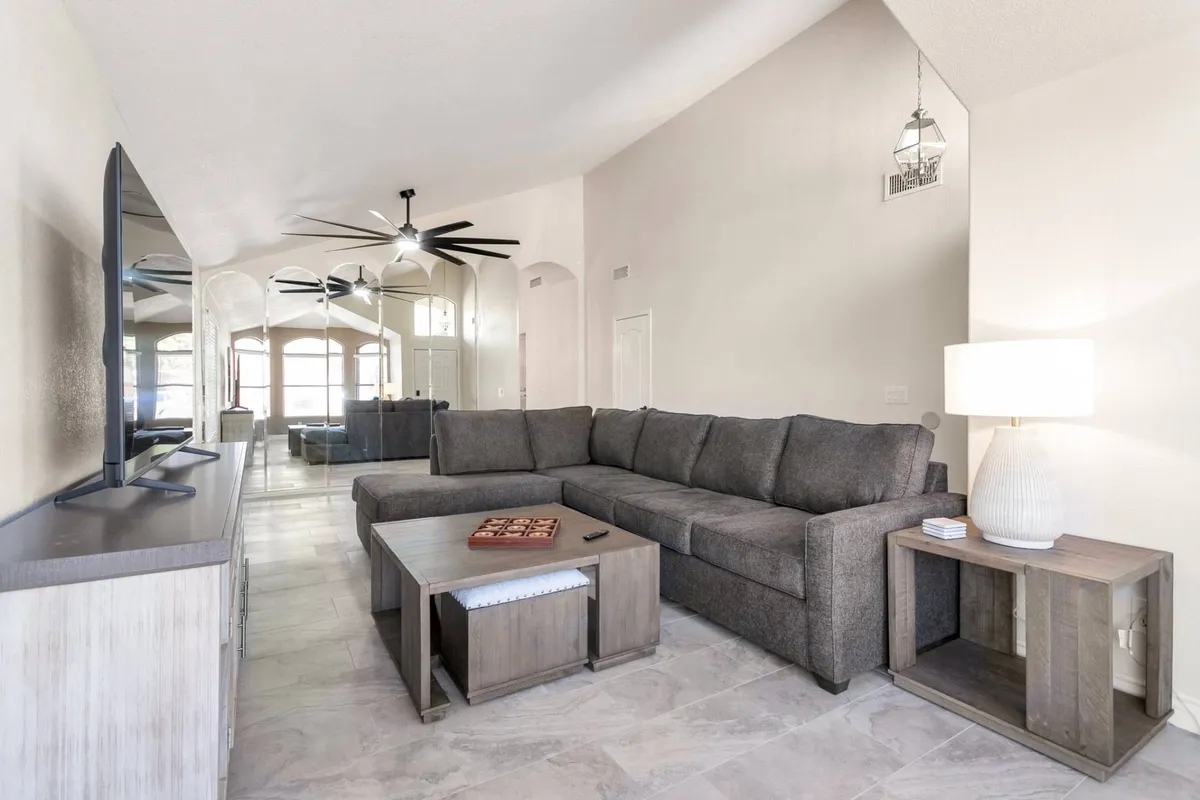 The image showcases a spacious living room featuring a large, plush sectional sofa arranged around a central coffee table, with a modern television mounted nearby. Elegant arched windows flood the space with natural light, while a ceiling fan overhead adds comfort. The setup invites guests to relax, socialize, enjoy movie nights, or gather for lively conversations.