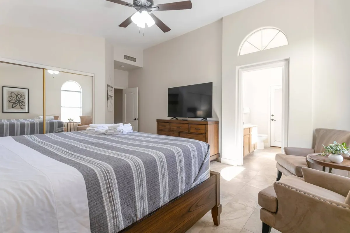 A spacious bedroom featuring a king-sized bed with plush linens, a cozy seating area for relaxation, a large television, and a mirror reflecting soft natural light. The atmosphere is comfortable and inviting, ideal for unwinding after a busy day.