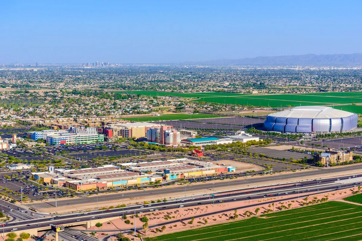 This wide-angle view captures a bustling urban landscape featuring a large stadium, a shopping center, expansive farmland, and a network of highways. The image conveys the energy and variety of activities available nearby, from sports events to shopping and exploring local parks.