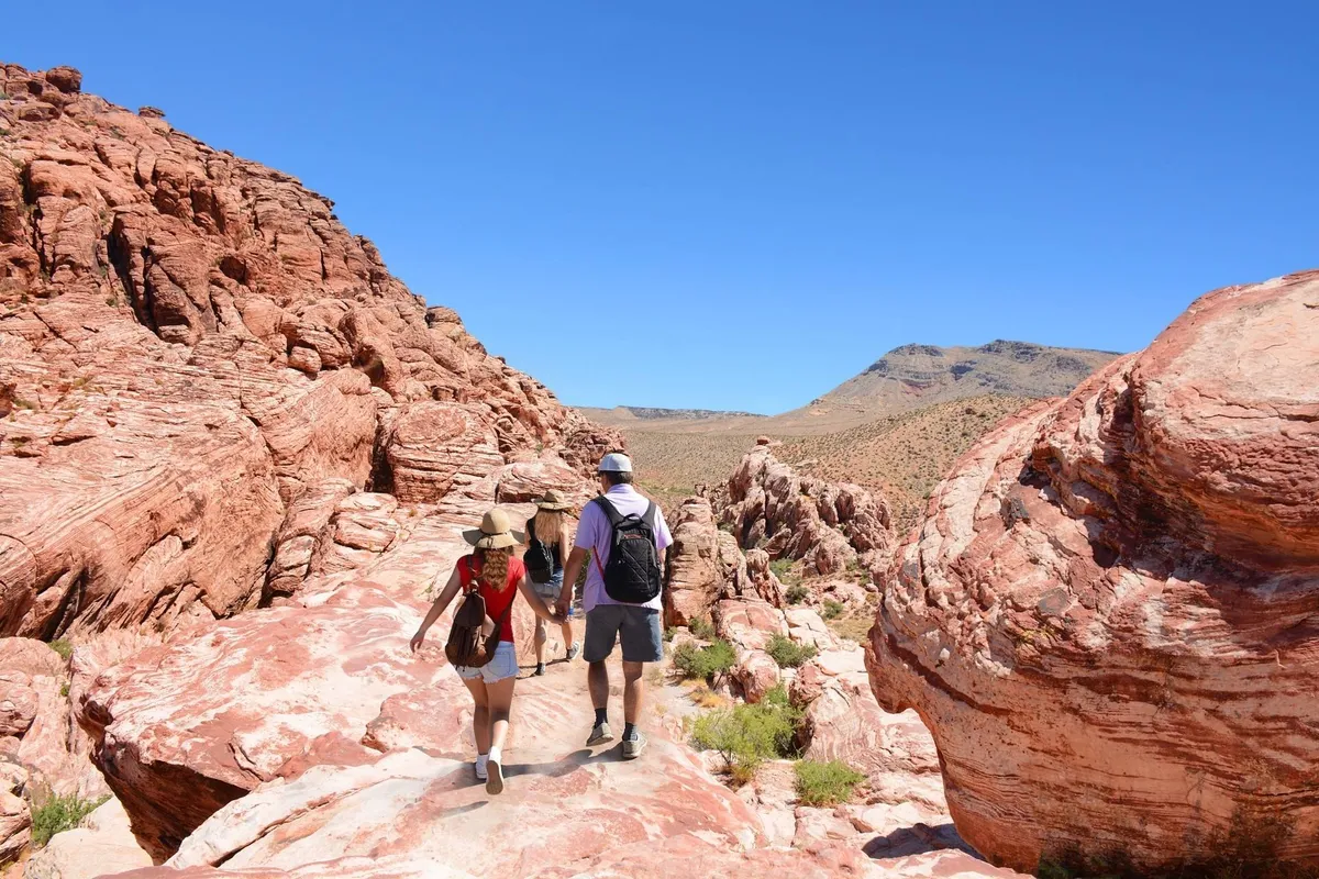 A couple is seen walking in front of dramatic red rock formations, with a wide blue sky overhead. The setting suggests a network of local trails or parks, perfect for outdoor activities and exploration.