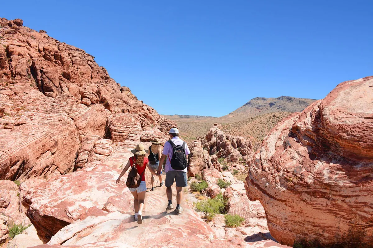 The image portrays a family-friendly adventure in the Arizona outdoors, with hikers exploring red rock formations under a bright, sunny sky. The dramatic mountain backdrop and natural landscape highlight the spirit of exploration and discovery.