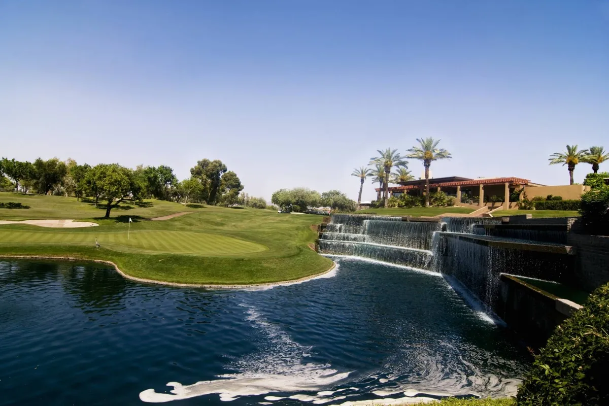 A vibrant exterior scene highlighting a lush golf course with a dramatic waterfall, pool area, palm trees, and a building in the background. The expansive setting offers plenty of space for outdoor gatherings and recreation.