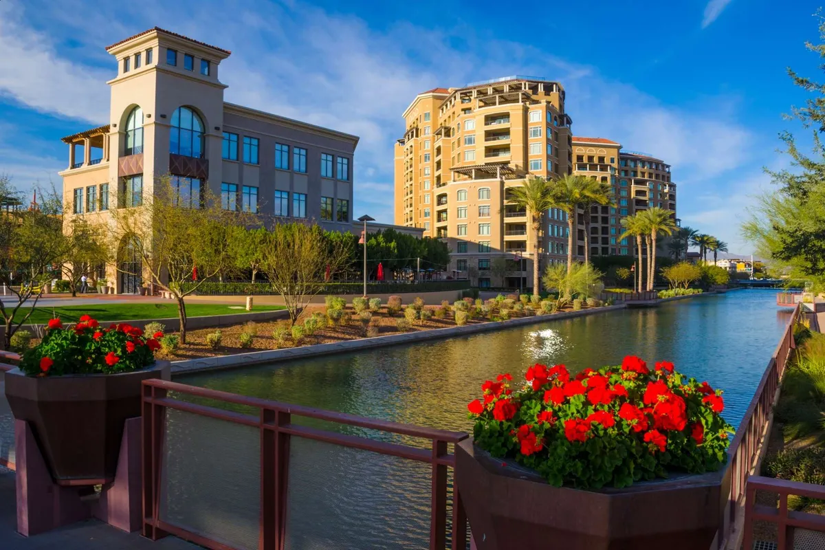 A vibrant, modern canal lined with palm trees, colorful flower pots, and contemporary buildings. People stroll along the water, enjoying the lively outdoor cafes and open spaces, suggesting local dining and networking opportunities.