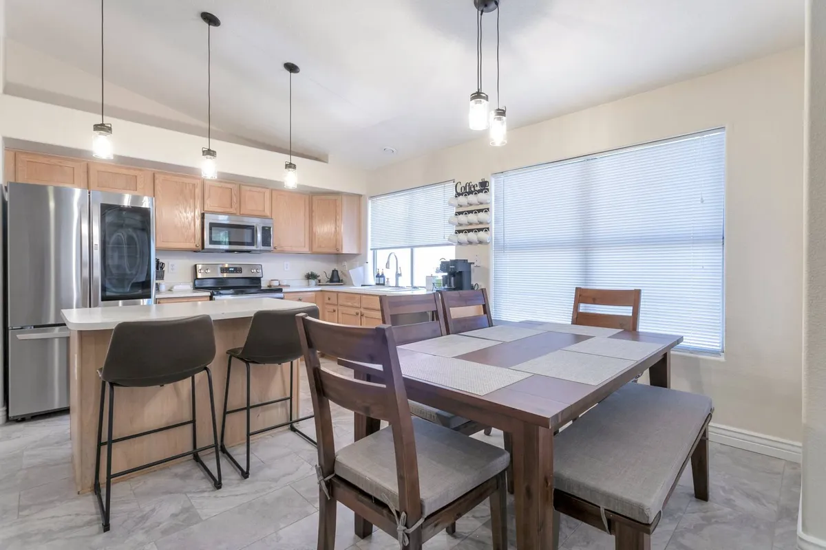 The dining area features a modern kitchen island, a spacious dining table, stylish pendant lighting, and a window with blinds that lets in natural light. The setup offers ample space for meals, board games, or casual chats, seamlessly connecting the kitchen and dining spaces.